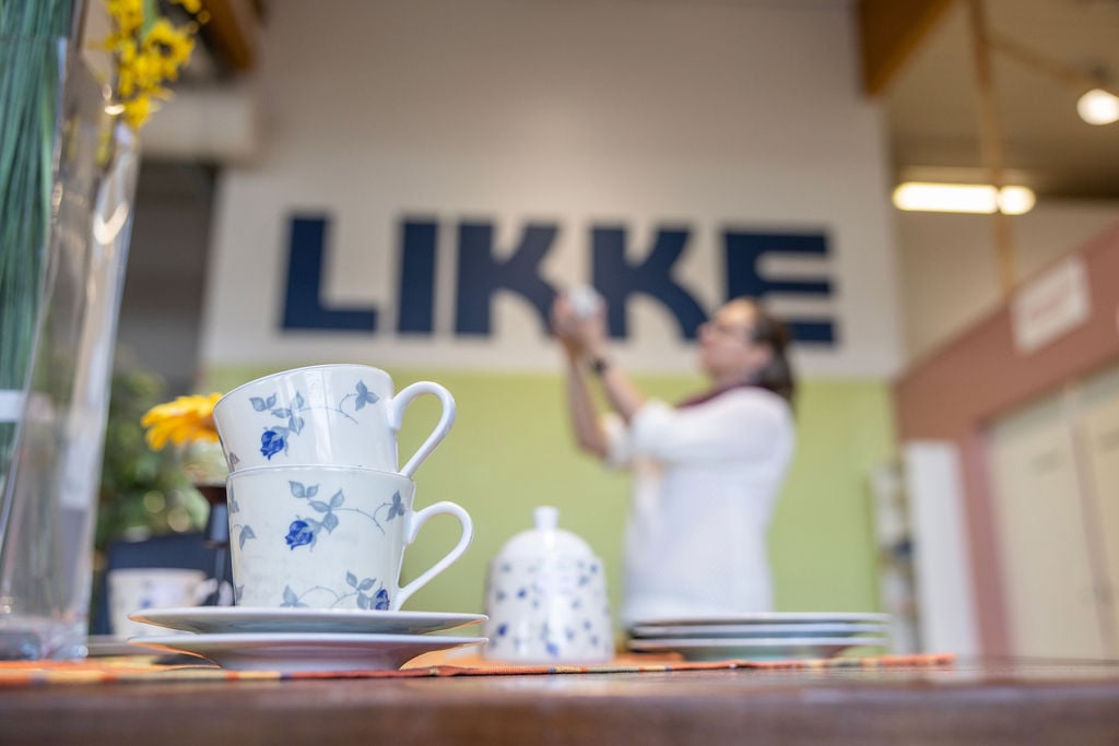 Kuvassa etualalla kahvikuppeja. Taustalla sumeana LIKKE-teksti ja nainen katselemassa esinettä käsissään.