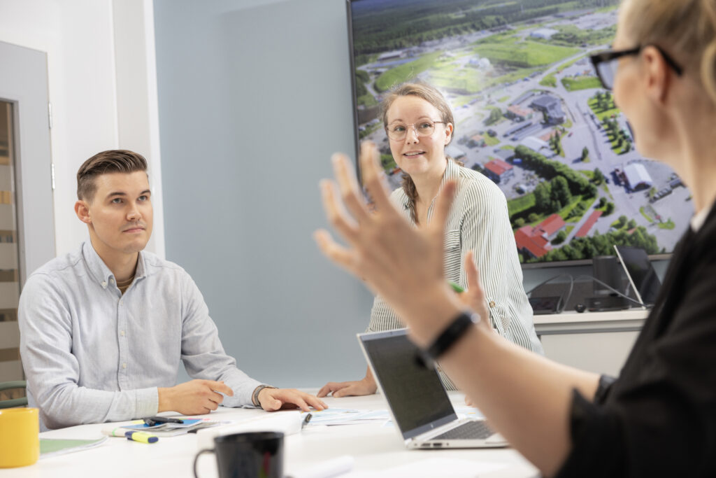 Mies ja kaksi naista neuvotteluhuoneen pöydän ääressä keskustelemassa innostuneena.