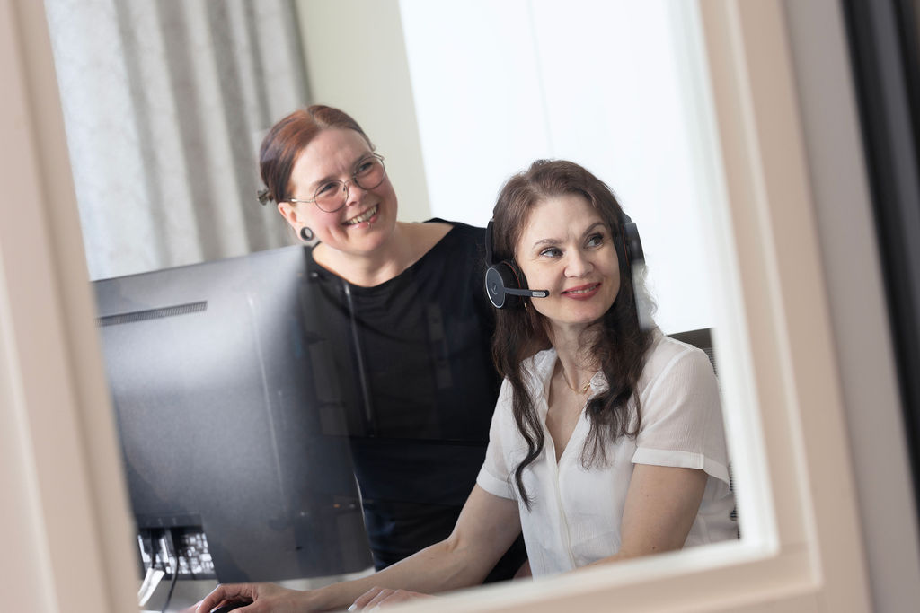 Kaksi hymyilevää naista tietokoneen ääressä, toinen seisoo, toisella on päässä kuulokkeet mikrofonilla.