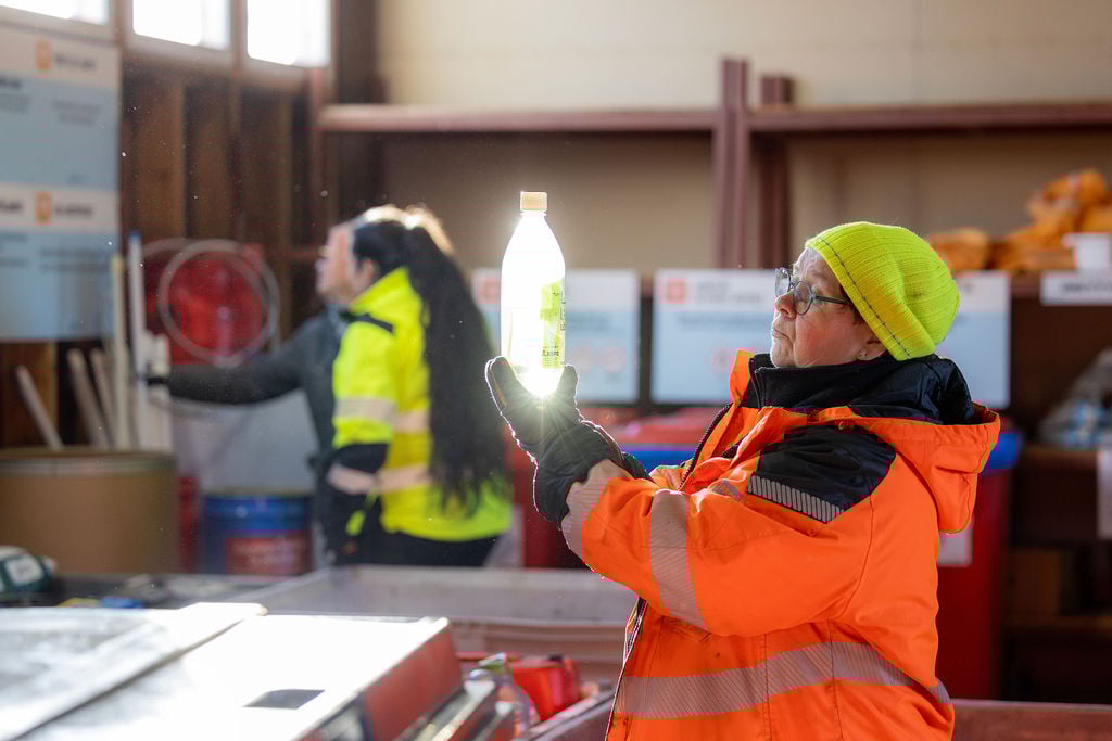 Nainen tutkii kädessään olevaa pulloa jäteasemalla. Pulloon osuu auringonvalo ja se kimaltaa.