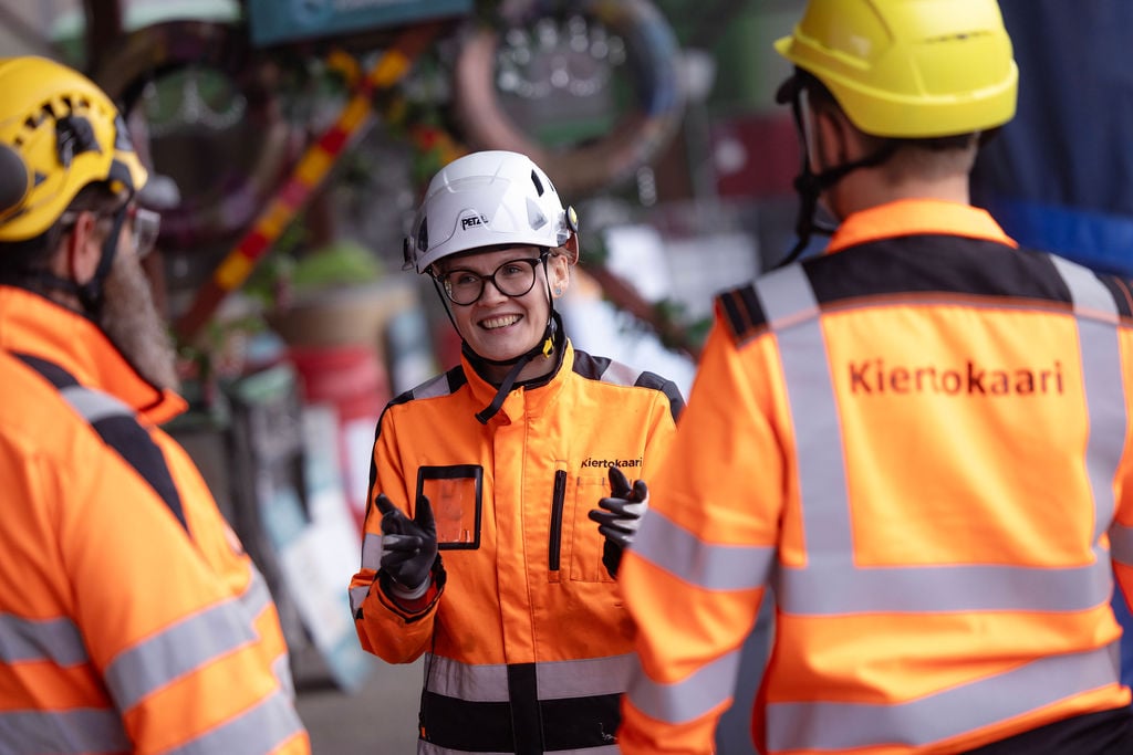hymyilevä työasuinen nainen juttelee kahden työasuisen miehen kanssa, jotka selin kameraan.
