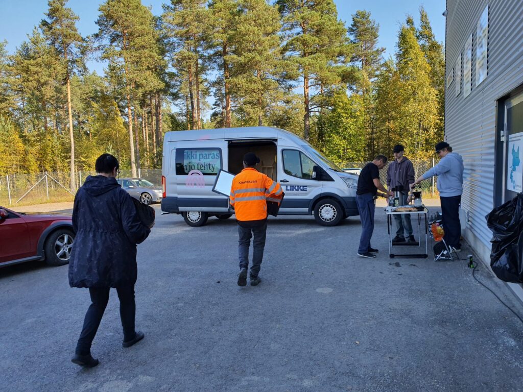 Asiakas tuo Liken keräysautoon (pakettiauto) tavaraa, kuvassa näkyy kiertokaaren henkilökuntaa kantamassa peiliä sekä ihmisiä ottamassa kahvia kahvipisteellä. 