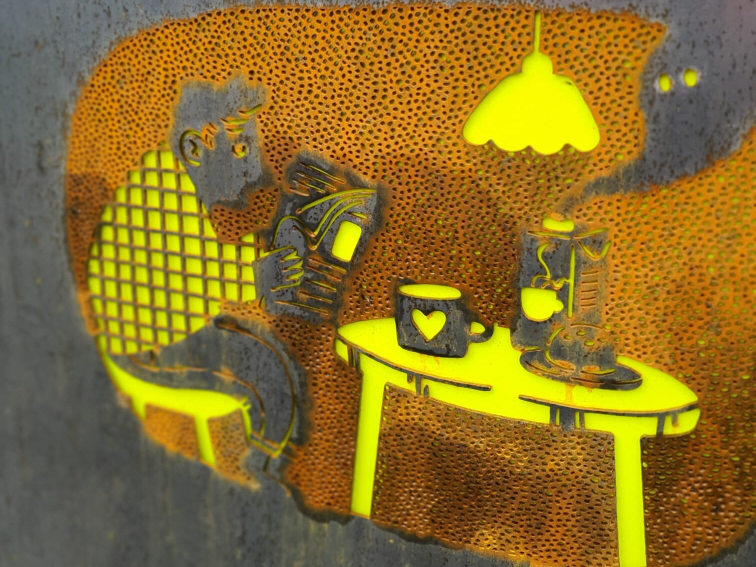 Jäteastian kyljessä oleva taideteos, jossa mies lukee lehteä kahvipöydässä. Kuva muodostuu pienistä teräspohjaan leikatuista rei'istä.