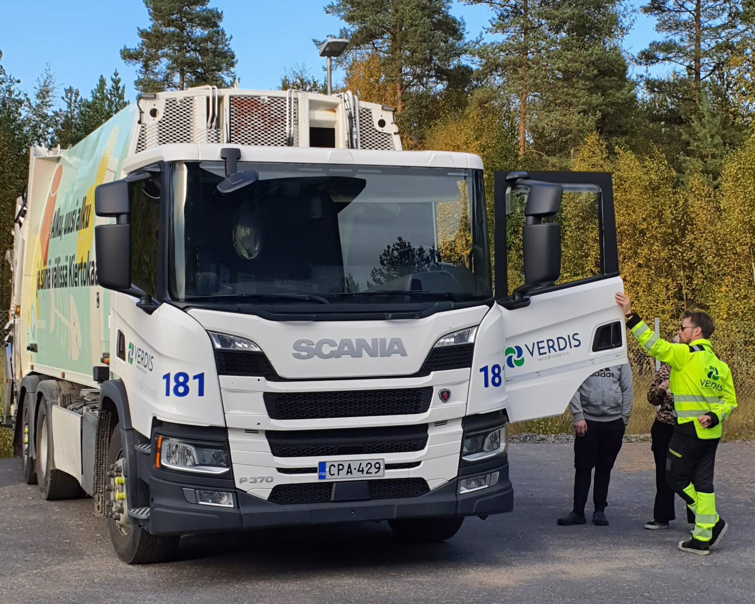 Jäteauto pysäköitynä. Kuljettaja pitää ovea auki ja autoon on tutustumassa yksi vierailija.