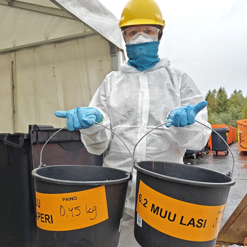 Valkoiseen suojahaalariin pukeutunut opiskelija pitää käsissään koostumustutkimuksen ämpäreitä. 