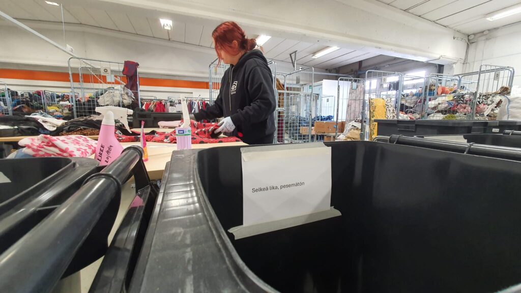 Nainen lajittelee tekstiilejä.. Etualalla jätastia, jossa lukee: selkeä lika, pesemätön.